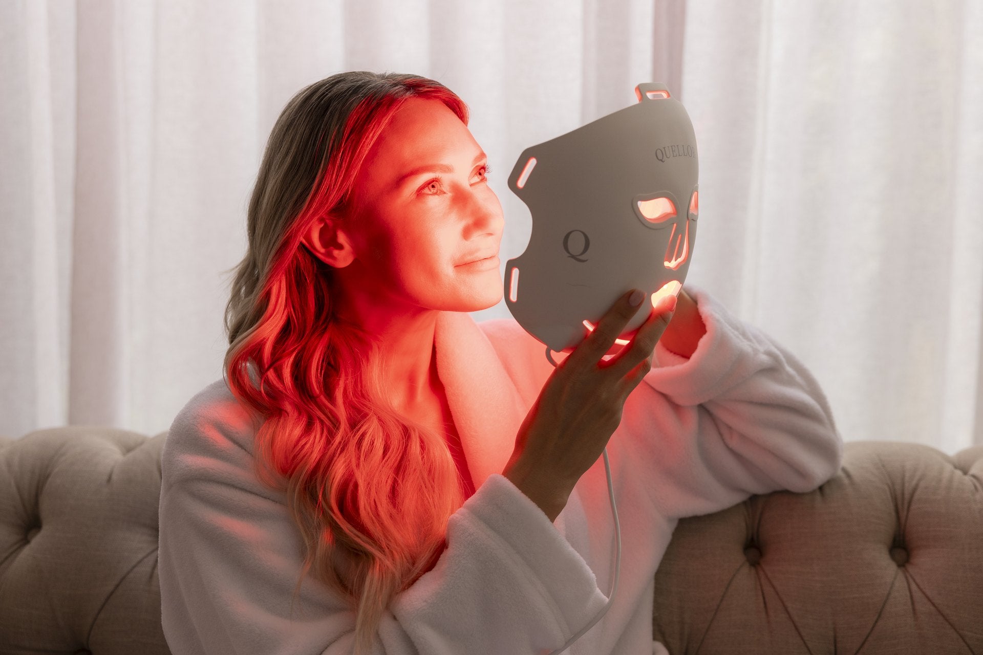 Woman holding an illuminated Quellor LED mask beside her face during a relaxed at-home treatment