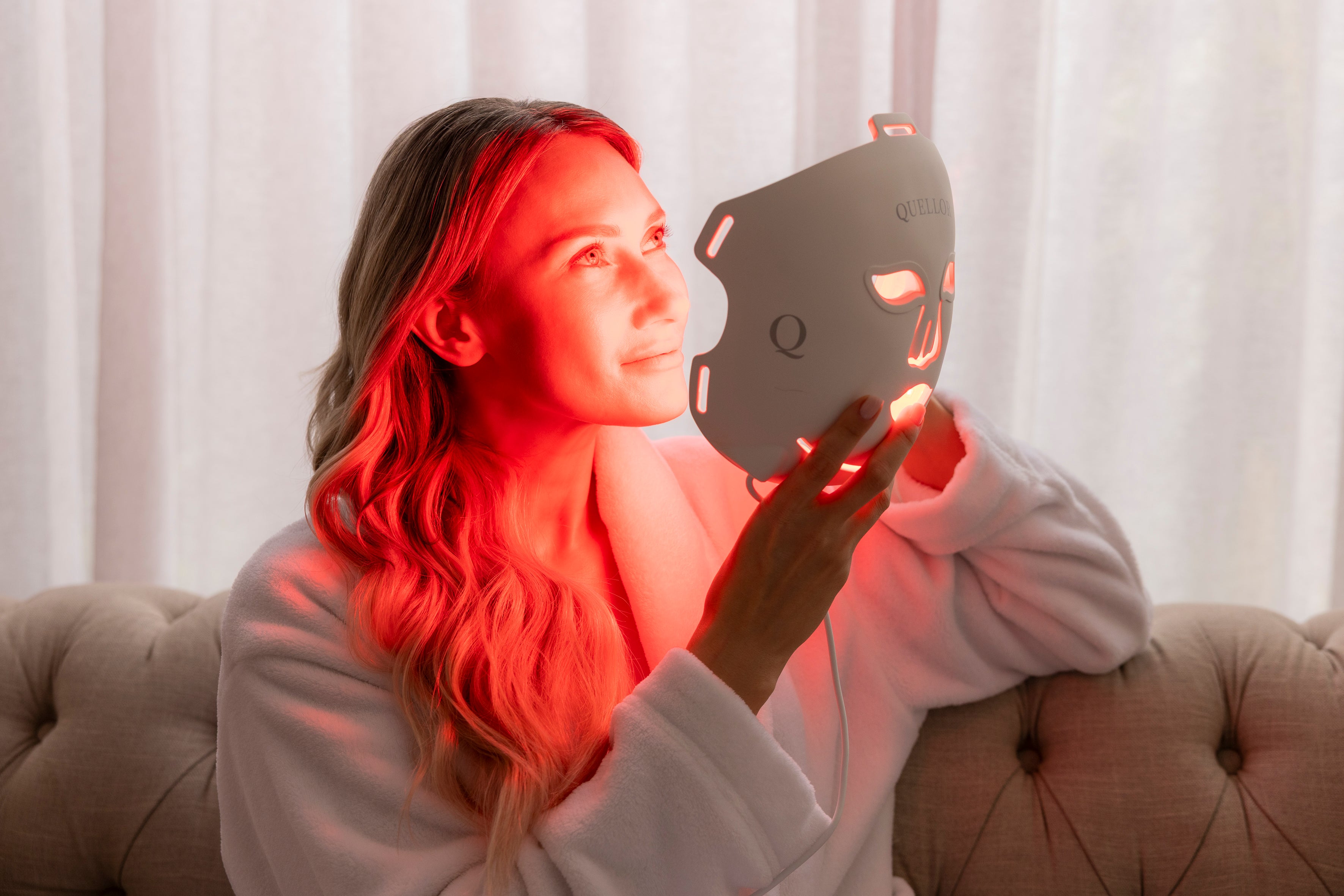 Woman holding the illuminated Quellor LED Light Therapy Mask during a relaxed at-home skincare session.