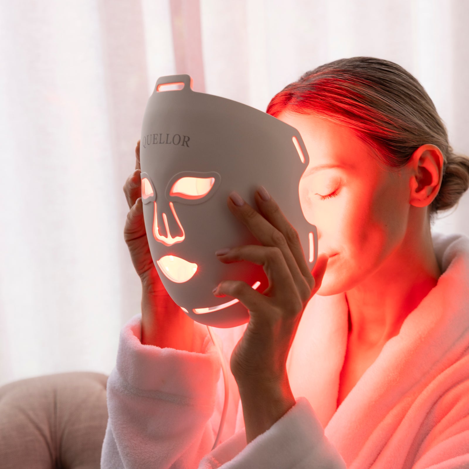 Woman holding the illuminated Quellor LED Light Therapy Mask near her face.